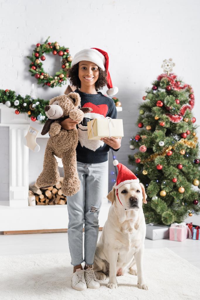 Веселая африканская американка с подарочной коробкой и плюшевым мишкой рядом с собакой-лабрадором и рождественской ёлкой  - Фото, изображение
