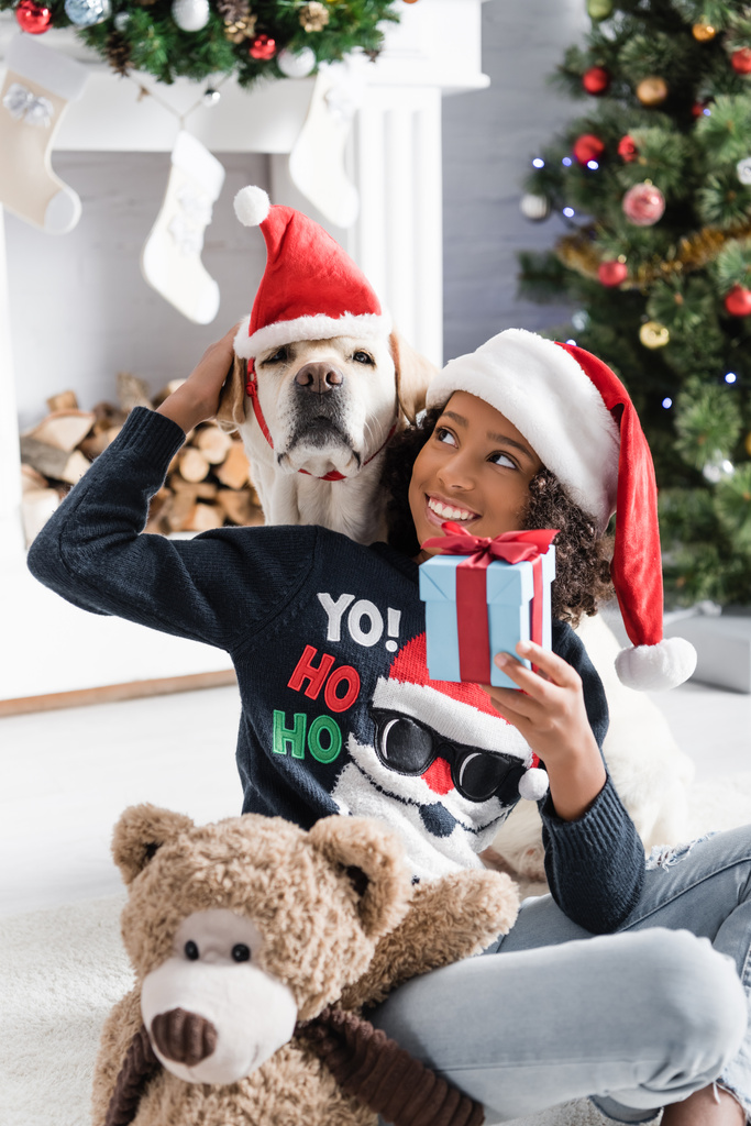 Gülümseyen Afrikalı Amerikalı kız, Labrador 'u okşarken hediye kutusu ve bulanık arka planda oyuncak ayıyla yerde oturuyor. - Fotoğraf, Görsel