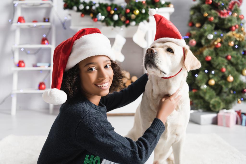 felice afro americano ragazza coccole labrador in cappello di Babbo Natale vicino albero di Natale su sfondo sfocato - Foto, immagini
