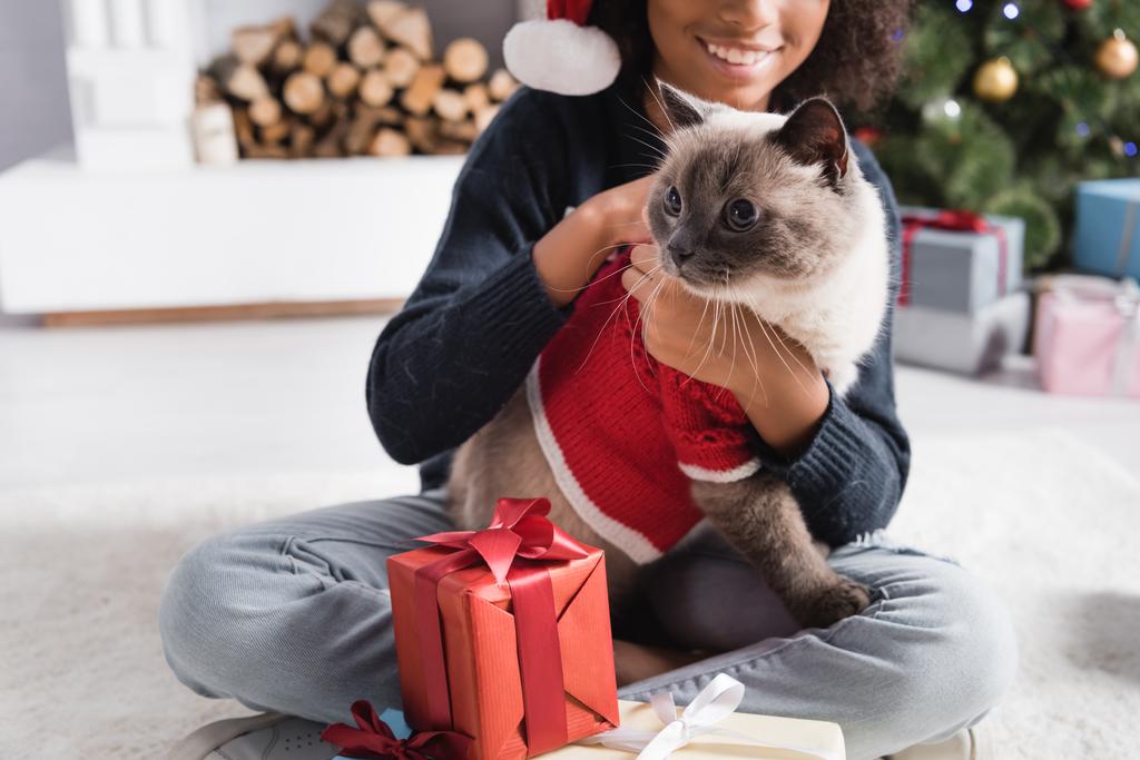 vue recadrée de fille afro-américaine câlinant chat tout en étant assis près de boîtes-cadeaux sur fond flou - Photo, image