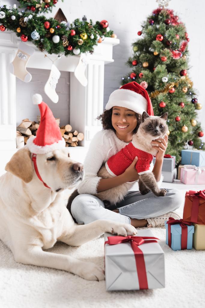Neşeli Afro-Amerikalı kız elinde tüylü kediyle Noel Baba şapkalı, bulanık ön planda hediye kutusuna dokunuyor. - Fotoğraf, Görsel