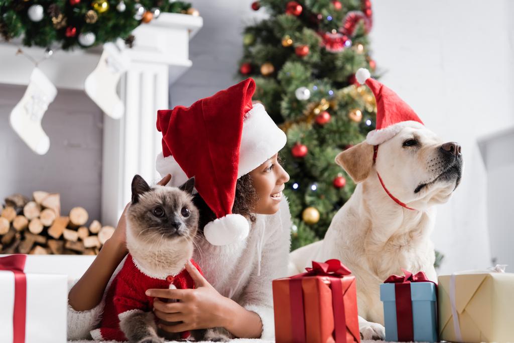 joyeuse fille afro-américaine regardant loin près de labrador, chat, boîtes-cadeaux et arbre de Noël sur fond flou - Photo, image