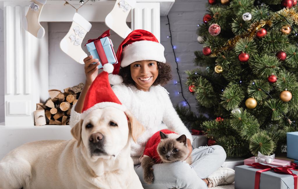 radostný africký americký dívka drží vánoční dárek v blízkosti labrador pes na rozmazané popředí   - Fotografie, Obrázek