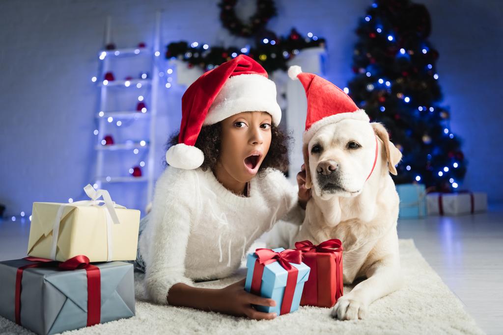 izgatott afro-amerikai lány fekvő padlón közelében labrador kutya és karácsonyi ajándékok elmosódott háttér - Fotó, kép