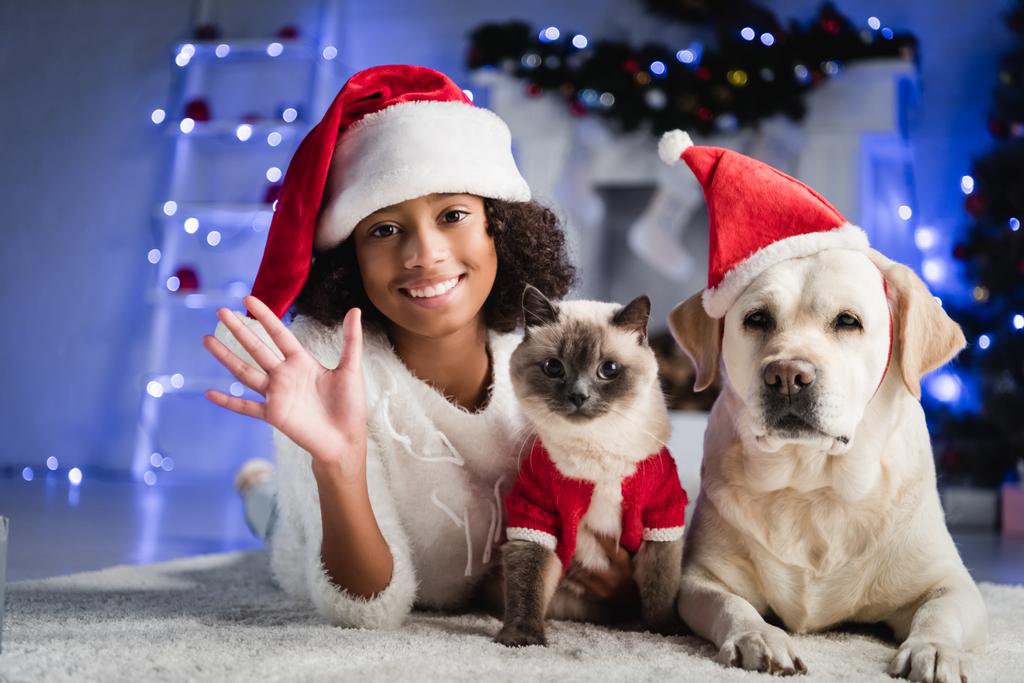 Heureuse fille afro-américaine avec main agitant couchée sur le sol près du chat et du labrador dans le chapeau de Père Noël sur fond flou - Photo, image
