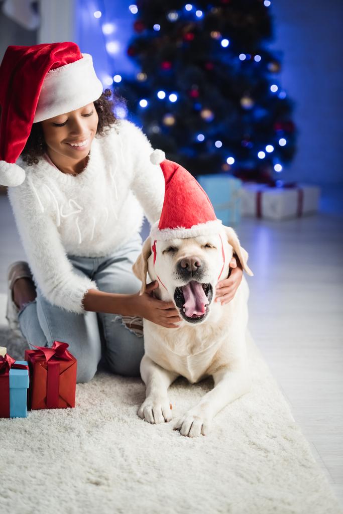 Smiling african american girl putting santa hat on yawning retriever, sitting on rug near presents on blurred background - Photo, Image