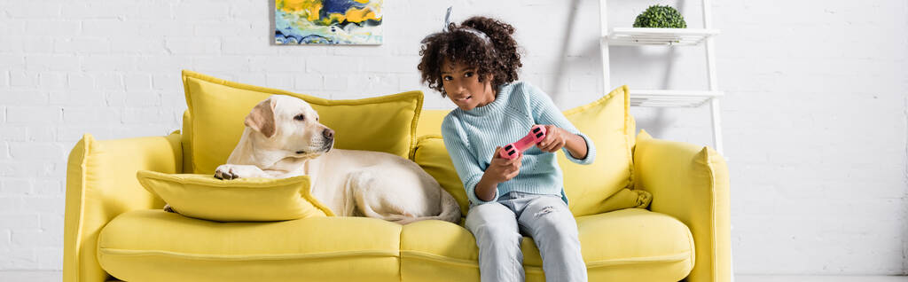 KYIV, UKRAINE - OCTOBER 02, 2020: Focused girl playing with joystick, while sitting near labrador on sofa at home, banner - Photo, Image