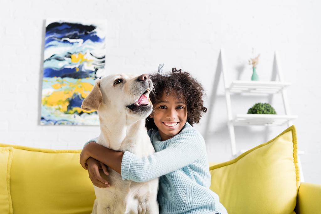 Šťastná kudrnatá dívka objímající labrador štěkání, zatímco sedí na žluté pohovce doma - Fotografie, Obrázek