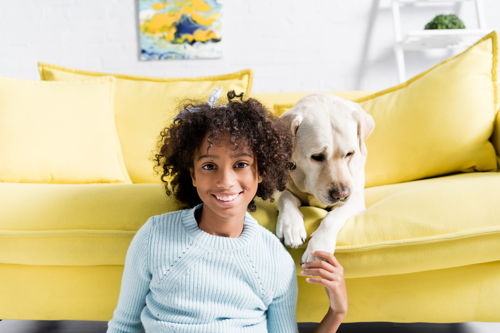 Улыбающаяся девушка касается лабрадора лапы, глядя в камеру и сидя рядом с диваном, дома, на размытом фоне - Фото, изображение