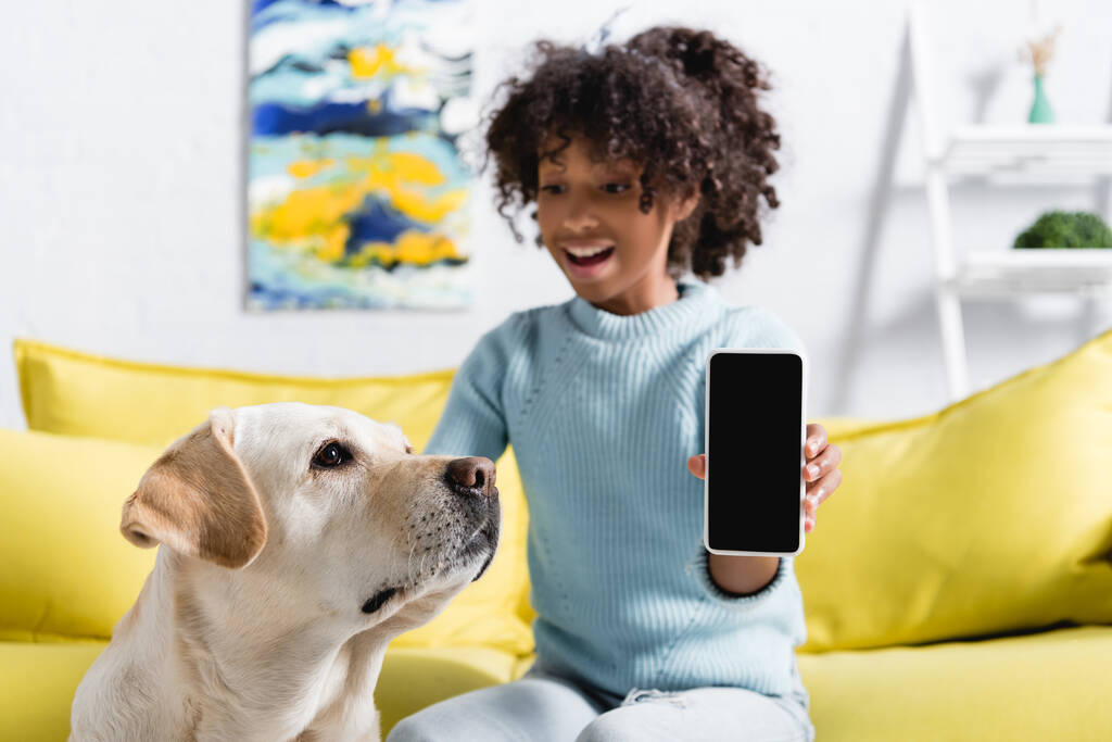 Retriever zitten in de buurt glimlachend Afrikaans Amerikaans meisje tonen smartphone met leeg scherm op wazig achtergrond - Foto, afbeelding
