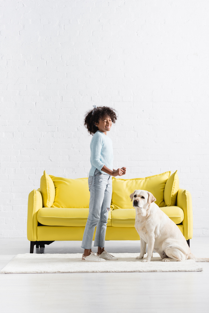 Лабрадор сидить поруч з афро-американською дівчинкою, яка сміється, стоячи на білому килимі вдома. - Фото, зображення