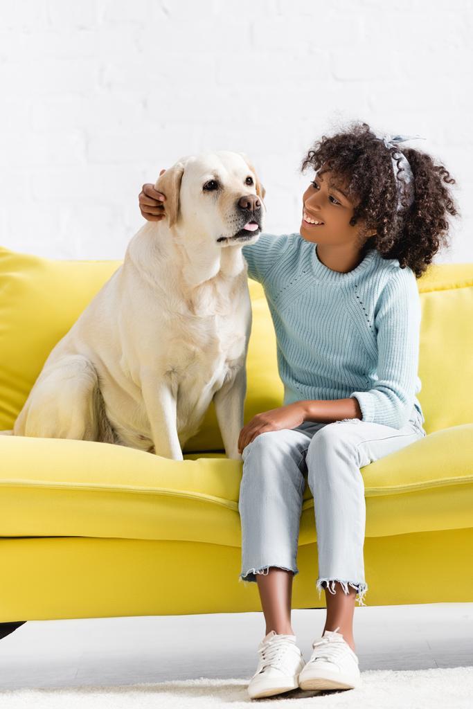 Smiling african american girl embracing and looking at retriever, while sitting on yellow sofa at home - Foto, afbeelding
