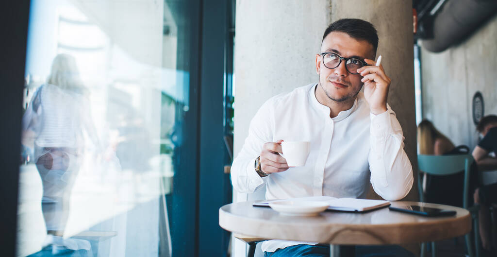 Przemyślany młody mężczyzna w białej koszuli i okularach pijący kawę podczas pracy z notatkami w nowoczesnej kawiarni i odwracając wzrok - Zdjęcie, obraz