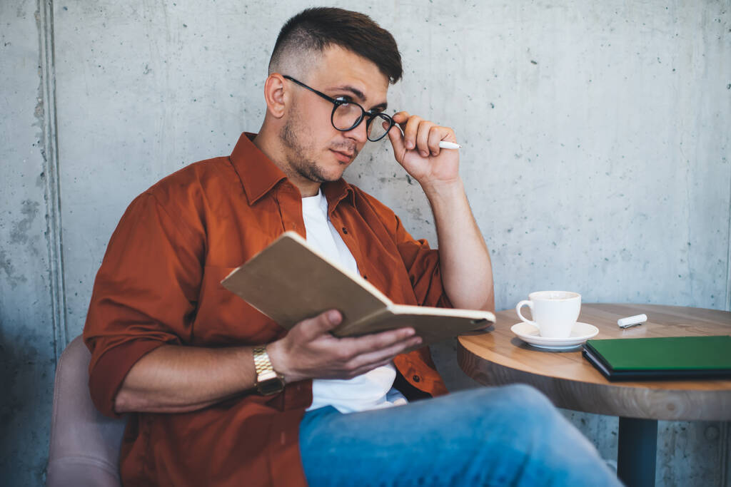 Pensive mladý muž na volné noze v brýlích nosí červenou košili a džíny myšlení a dotýkání brýlí při psaní poznámek v denním plánovači a sedí u stolu s šálkem kávy v blízkosti šedé betonové stěny kreativní kavárny - Fotografie, Obrázek