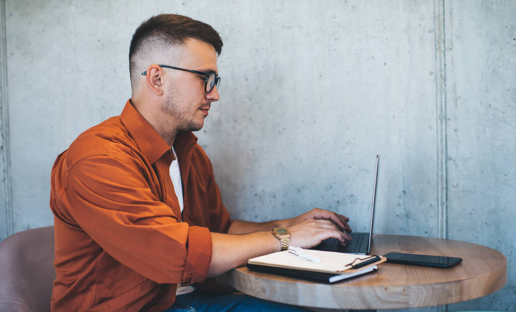 Вид сбоку на ориентированного молодого человека в джинсах оранжевой рубашки и очках, работающего над проектом на ноутбуке, сидящего за столом в кафетерии - Фото, изображение