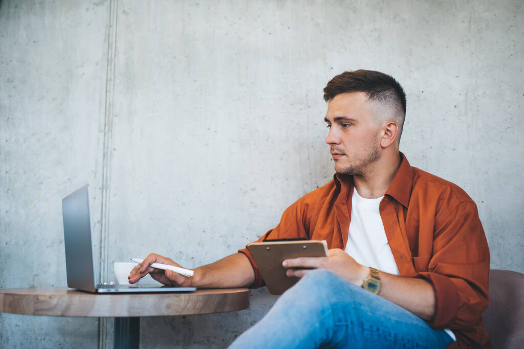 Молодой мужчина с бородой и короткой стрижкой в повседневной одежде, сидя за столом и просматривая ноутбук во время работы над проектом - Фото, изображение