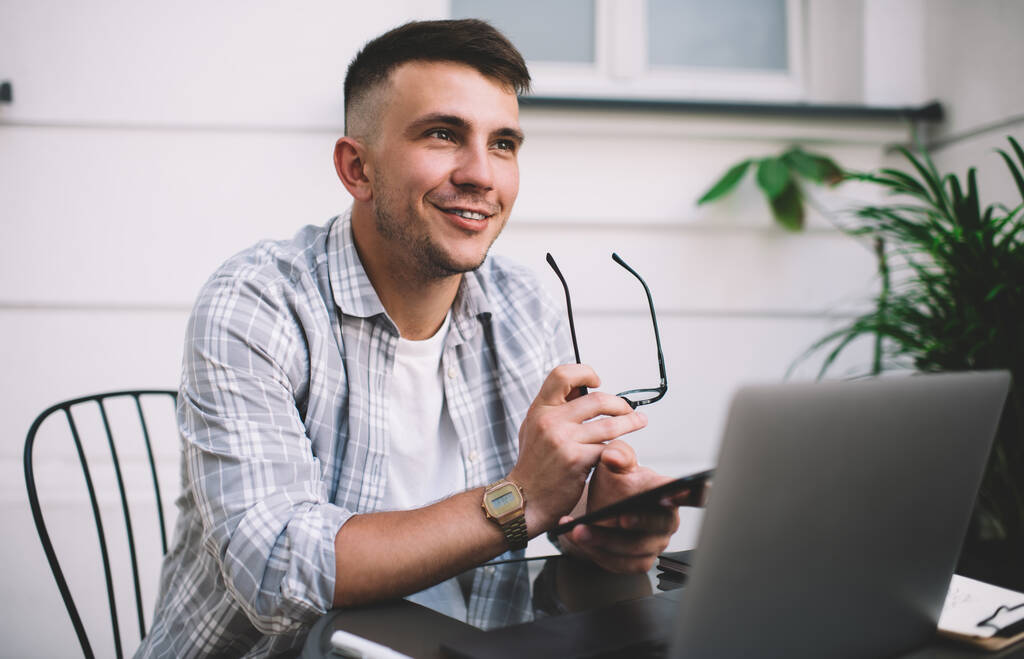 Homme joyeux en vêtements décontractés souriant et tenant des lunettes et un téléphone portable dans les mains tout en étant assis avec un ordinateur portable dans le café - Photo, image