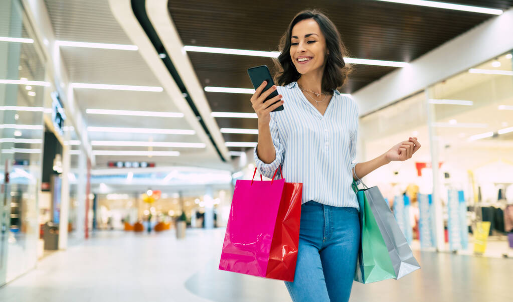 明るくカラフルなショッピングバッグで流行のスタイリッシュな服で現代的な豪華な若い笑顔の女性は、モールを歩いている間に彼女のスマートフォンを使用しています - 写真・画像