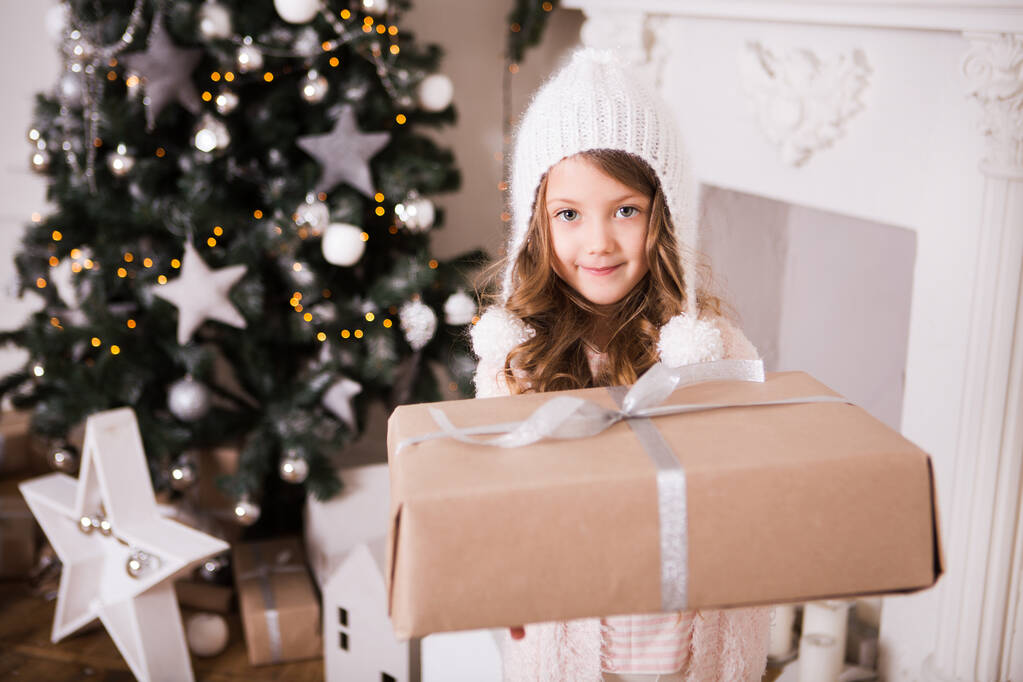 милая маленькая девочка в белой теплой зимней одежде и шляпе с подарочной коробкой позировать в украшенной Рождество комнате - Фото, изображение