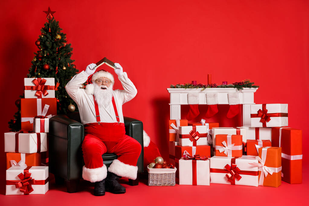 Porträt seiner er attraktive Fett übergewichtig funky kindisch fröhlich froh Weihnachtsmann sitzt im Sessel halten Buch wie Dach Spaß isoliert hell lebendigen Glanz lebendigen roten Farbhintergrund - Foto, Bild