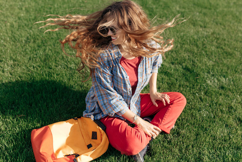 Χαρούμενη νεαρή ξανθιά hipster μαθήτρια με σακίδιο κάθεται στο γρασίδι στο πανεπιστήμιο. - Φωτογραφία, εικόνα