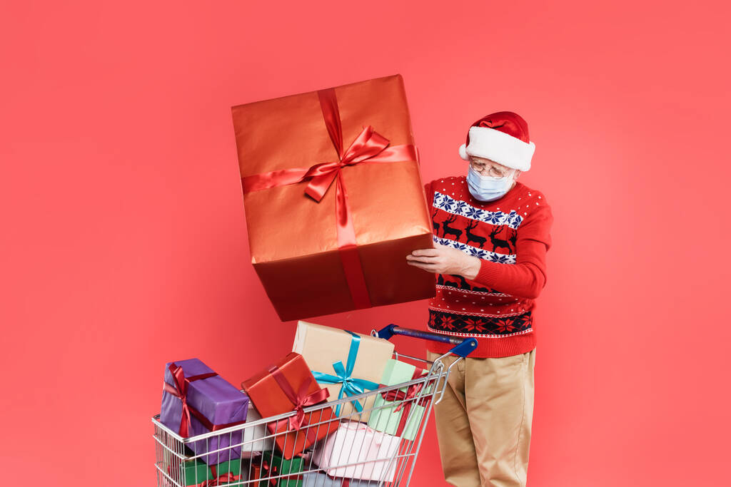 Elderly man in santa hat and medical mask holding gift near shopping cart with presents isolated on red  - Photo, Image