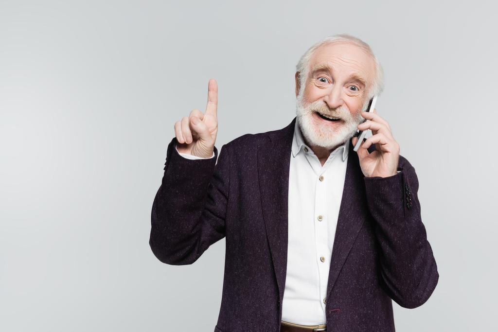 Hombre mayor positivo hablando en smartphone y señalando con el dedo aislado en gris  - Foto, Imagen