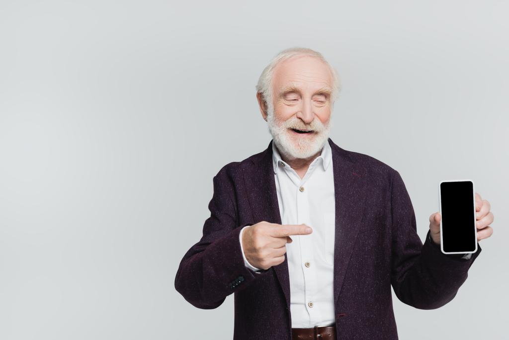 Hombre mayor sonriente apuntando con el dedo al teléfono inteligente con pantalla en blanco aislado en gris  - Foto, imagen