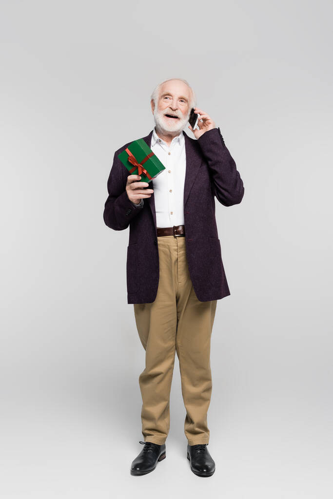 Позитивний старший чоловік розмовляє на смартфоні, тримаючи подарункову коробку на сірому фоні
  - Фото, зображення