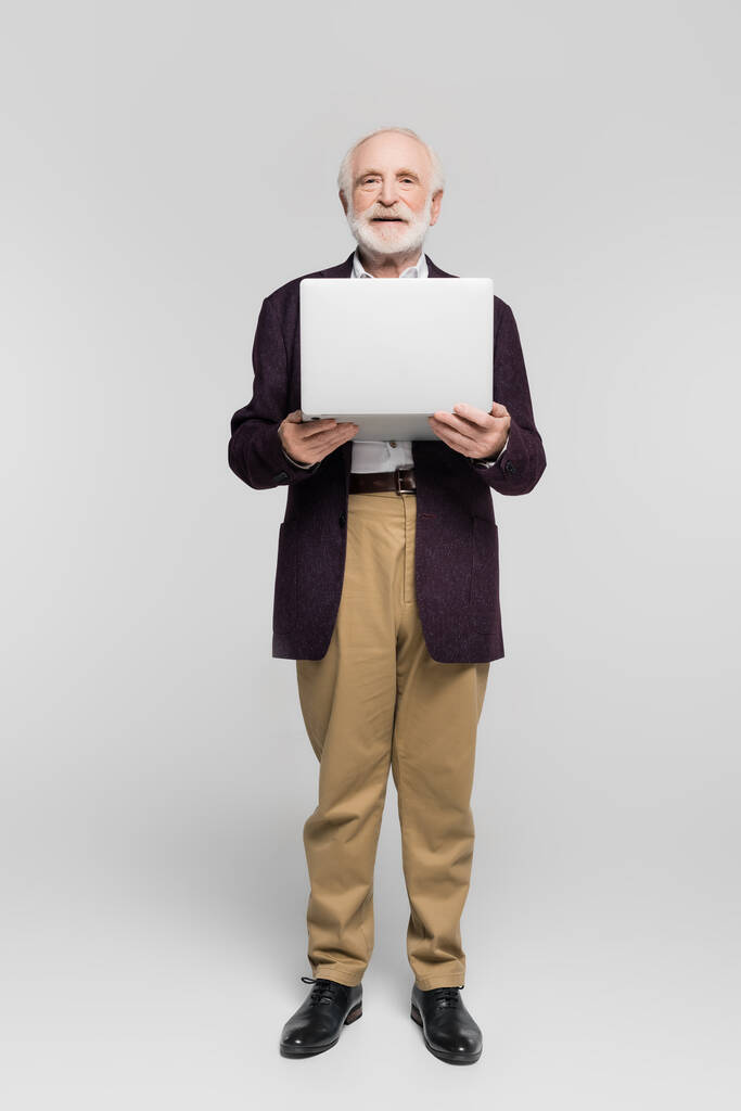 Senior Mann lächelt in die Kamera, während er Laptop auf grauem Hintergrund hält - Foto, Bild