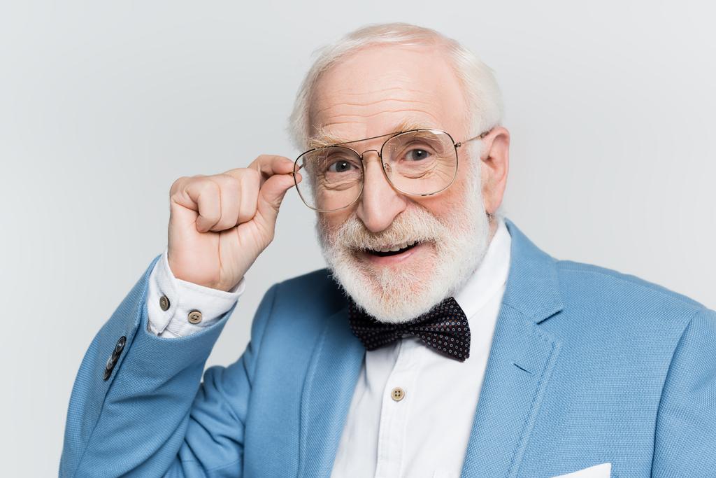 Uomo anziano positivo in giacca blu e papillon toccare occhiali isolati su grigio  - Foto, immagini