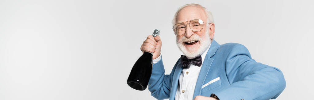 Χαμογελώντας ηλικιωμένος με παπιγιόν κρατώντας ένα μπουκάλι σαμπάνιας απομονωμένο στο γκρι πανό.  - Φωτογραφία, εικόνα