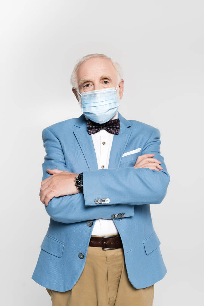 Hombre de pelo gris con máscara médica con brazos cruzados mirando a la cámara aislada en gris  - Foto, Imagen