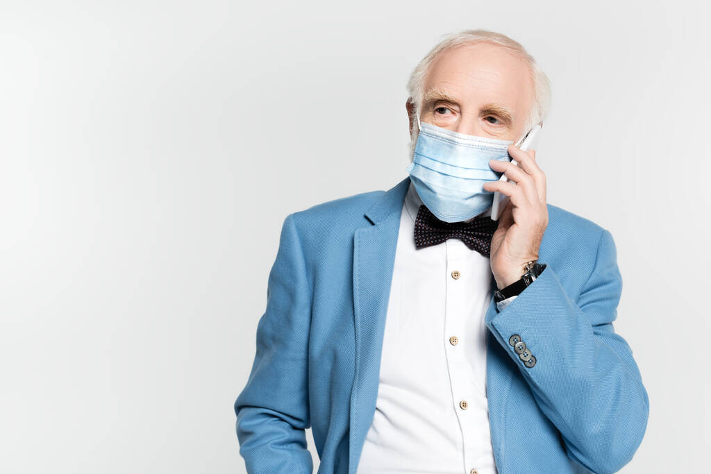 Hombre mayor en máscara médica hablando en el teléfono inteligente aislado en gris  - Foto, imagen