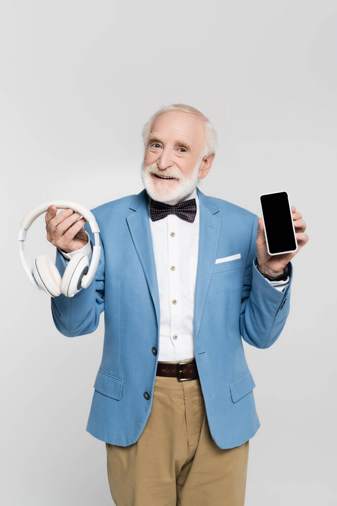 Θετικός ηλικιωμένος άνδρας με παπιγιόν και σακάκι που κρατά ακουστικά και smartphone με λευκή οθόνη απομονωμένη σε γκρι  - Φωτογραφία, εικόνα