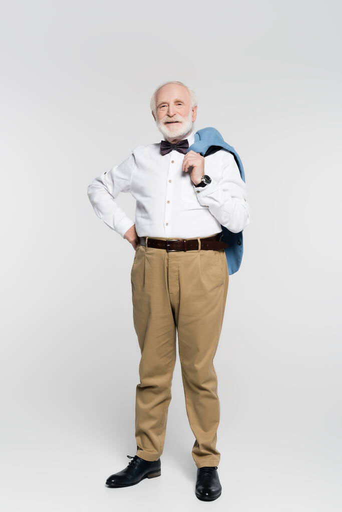Χαμογελώντας ηλικιωμένος άνδρας με το χέρι στο ισχίο κρατώντας σακάκι σε γκρι φόντο  - Φωτογραφία, εικόνα