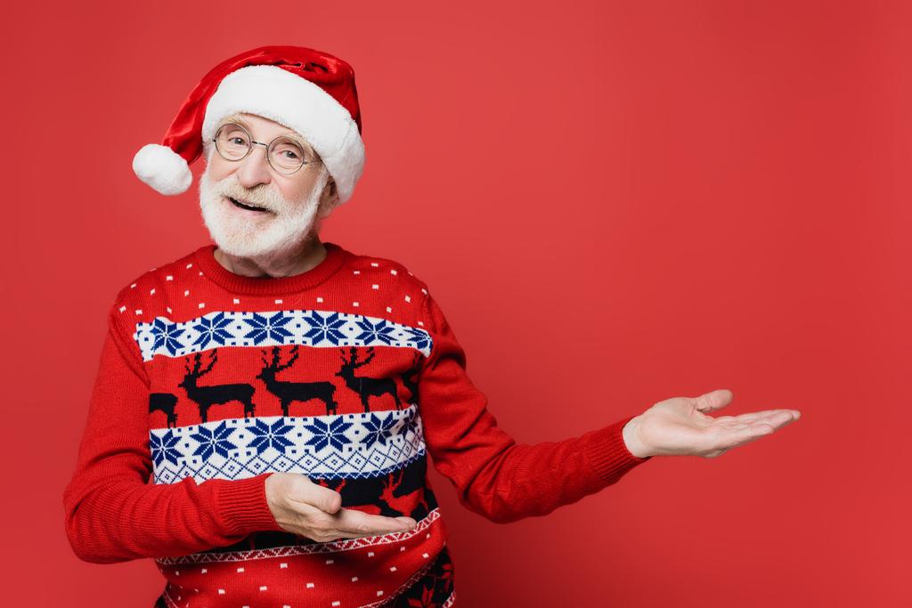 Homme âgé souriant en pull et chapeau de Père Noël pointant avec les mains sur fond rouge - Photo, image
