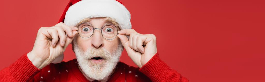 Здивований старший чоловік у капелюсі Санта та окулярах дивиться на камеру ізольовано на червоному, банер
  - Фото, зображення