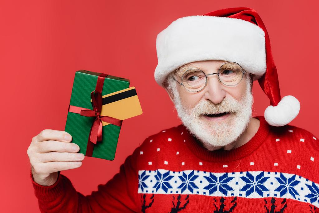 Idős férfi télapó kalapban és kötött pulóverben, ajándékdobozzal és hitelkártyával a kezében, amit piros alapon izoláltak.  - Fotó, kép
