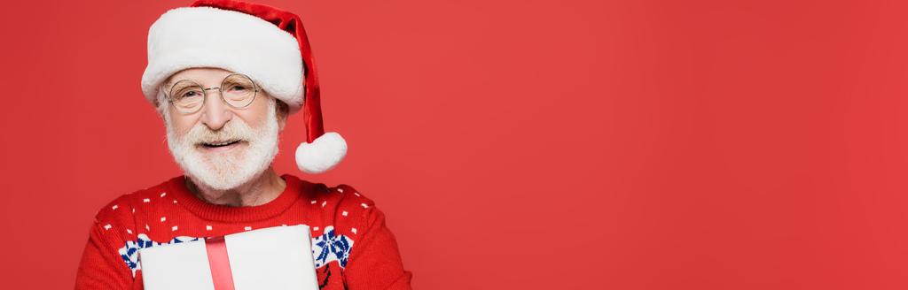Homme âgé souriant au chapeau de Père Noël tenant présent isolé sur rouge, bannière  - Photo, image