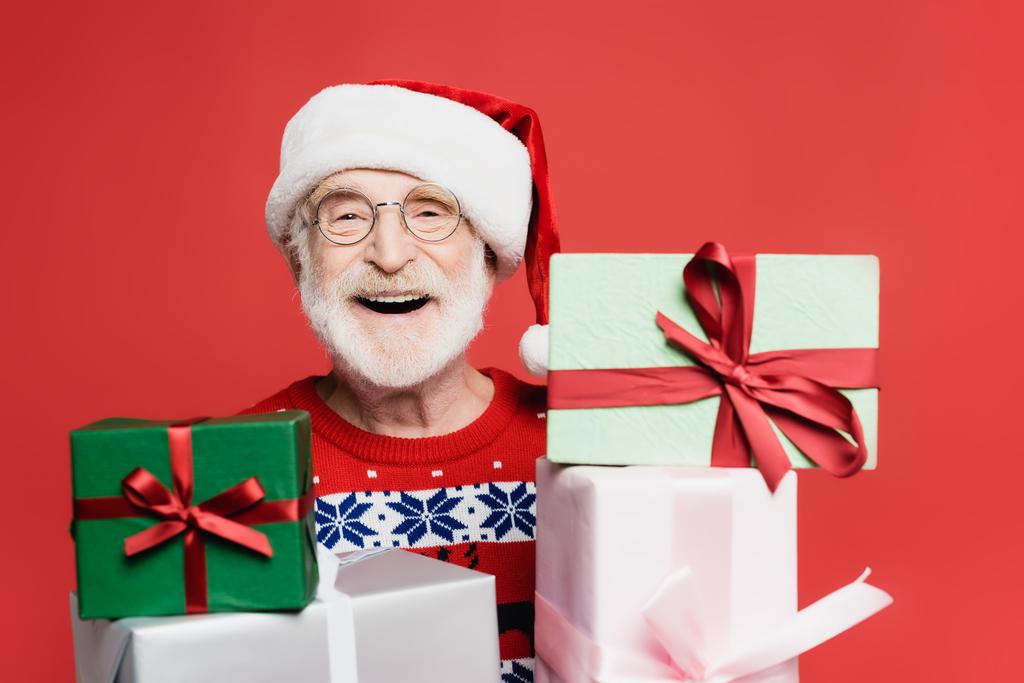 Χαρούμενος ηλικιωμένος άνδρας με καπέλο Σάντα κοιτάζοντας κάμερα κοντά σε δώρα σε θολή πρώτο πλάνο απομονωμένος σε κόκκινο.  - Φωτογραφία, εικόνα