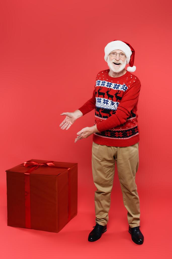 glimlachende senior man in santa hoed wijzend naar geschenken op rode achtergrond - Foto, afbeelding