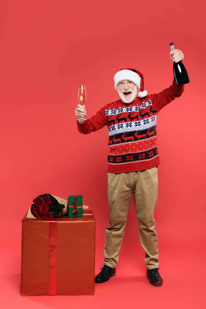 Веселий старший чоловік у капелюсі Санта тримає пляшку і келих шампанського біля подарунків і троянд на червоному тлі
 - Фото, зображення