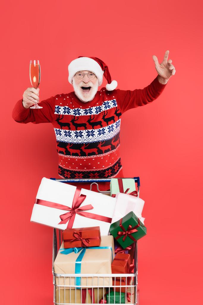 Vzrušený muž v Santa klobouku drží sklenici šampaňského v blízkosti nákupního košíku s dárky izolované na červené  - Fotografie, Obrázek