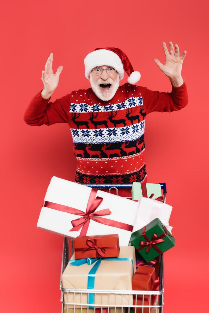 Schockierter älterer Mann mit Weihnachtsmütze blickt in die Kamera in der Nähe von Geschenken im Einkaufswagen  - Foto, Bild