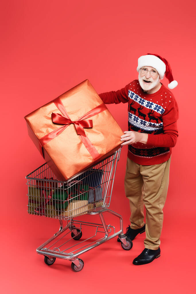 Smiling senior man in sweater and santa hat holding present near shopping cart on red background - Photo, Image