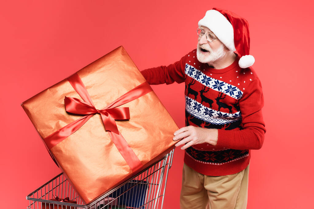 Positive senior man in santa hat and sweater holding gift with bow near shopping cart isolated on red  - Photo, Image