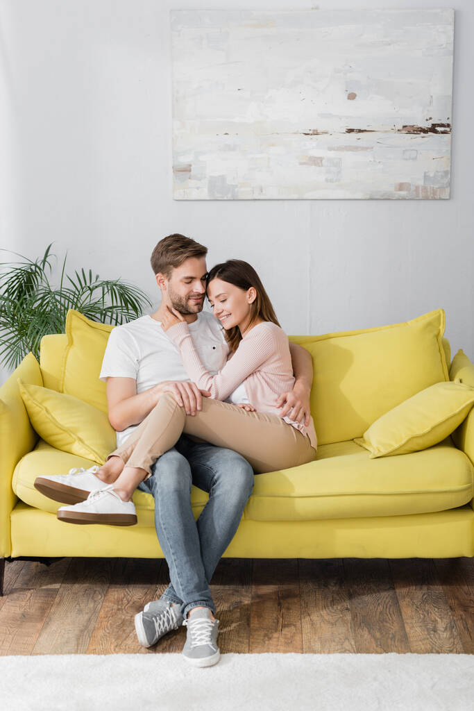 wesoły mężczyzna i kobieta przytulanie siedząc na kanapie w salonie  - Zdjęcie, obraz