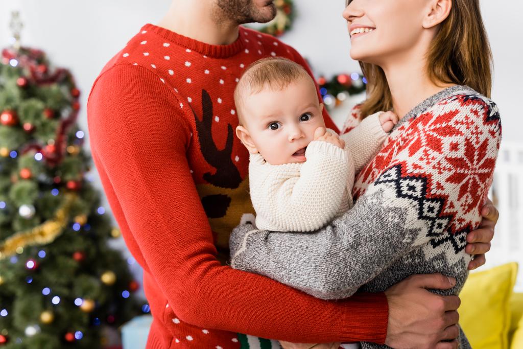 niño en brazos de la madre cerca del padre y borrosa árbol de Navidad en el fondo - Foto, Imagen