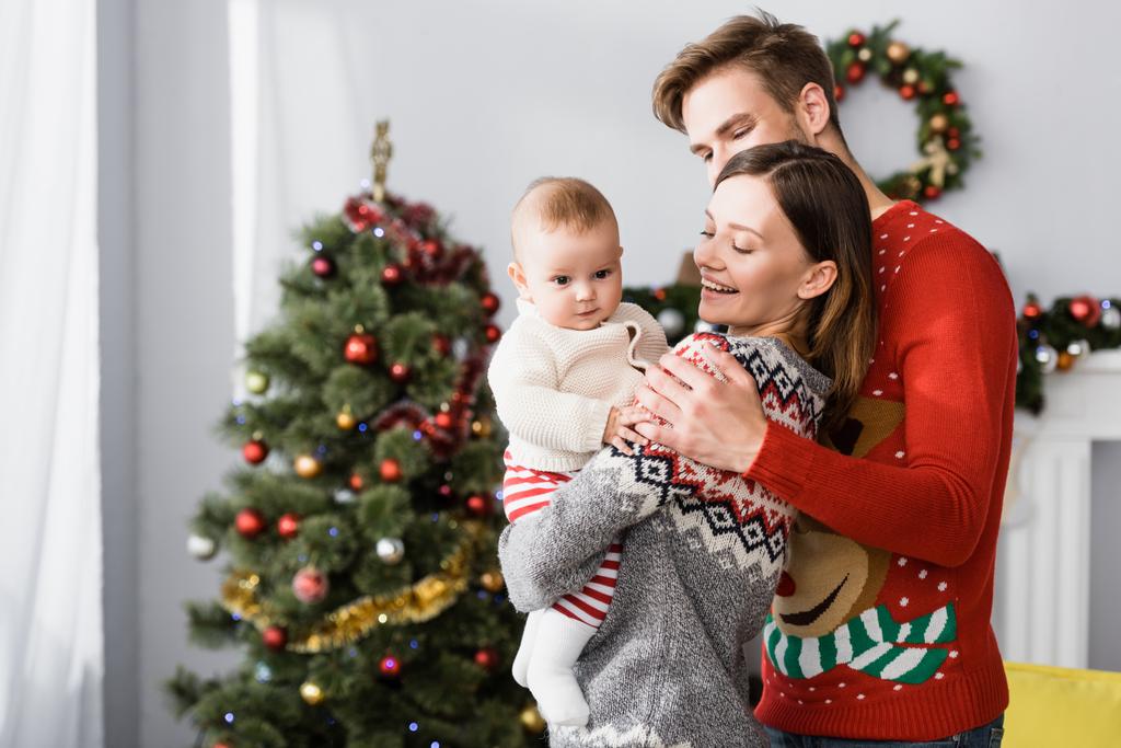 happy parents holding in arms baby boy near decorated christmas tree on blurred background - Photo, Image